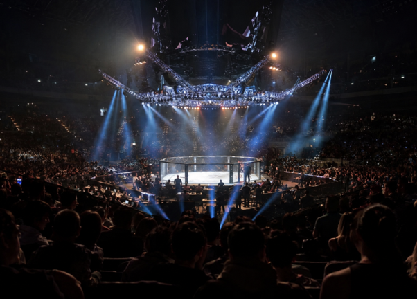 Packed MMA arena with a brightly lit octagonal cage awaiting the fighters.