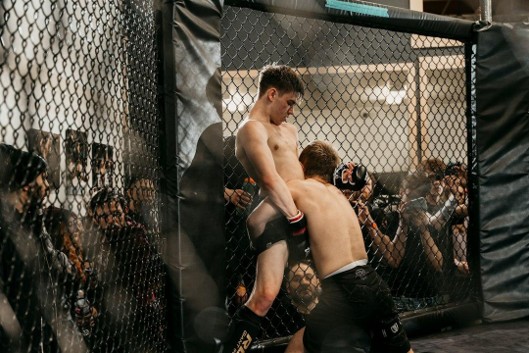 Two fighters clinching against the cage wall as spectators watch